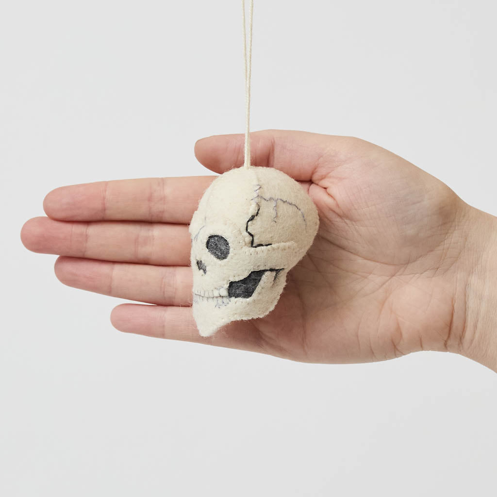 A handmade felted and embroidered Human Skull Ornament featuring embroidered bone, teeth, and crack details for a delightfully spooky addition to Halloween décor.