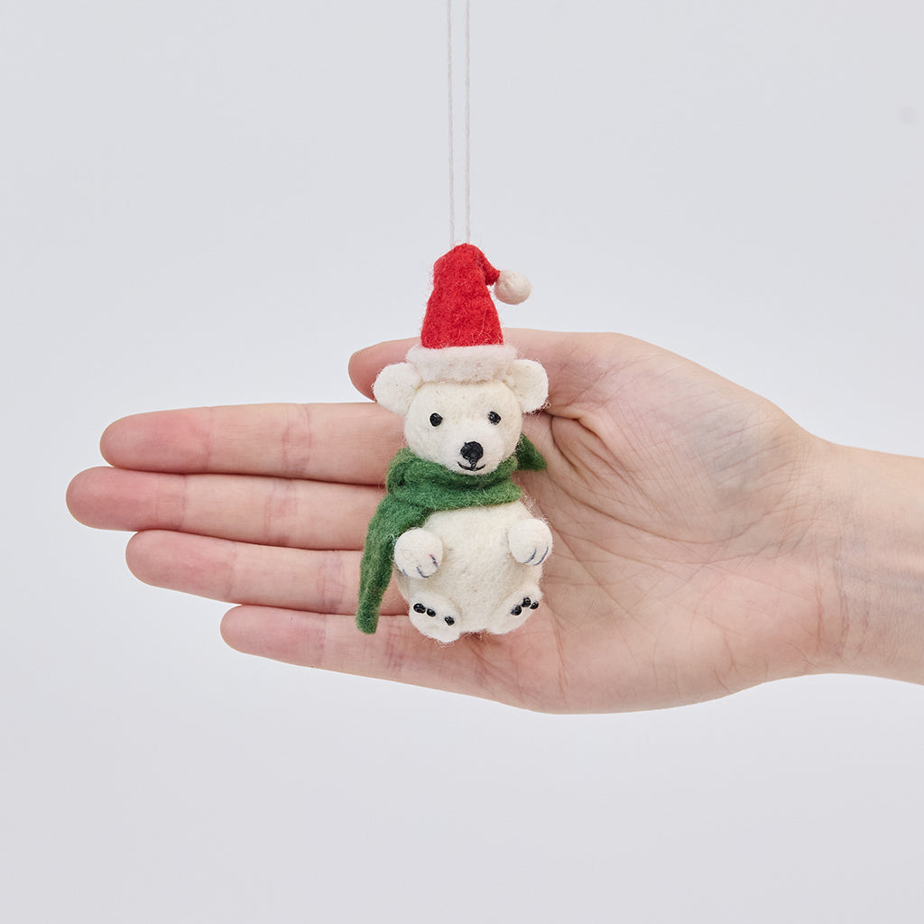 Handmade felt Little Drop Polar Bear ornament from Craftspring held in front of a hand to show its size—featuring a Santa hat and green scarf with detailed stitching, ethically crafted by artisans using sustainable wool.