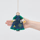 Craftspring’s Marie Curie ornament held in front of a hand to show its size—handmade from sustainable wool with intricate beaded and embroidered details including her holding two Nobel Prizes and a flask of radium, ethically crafted by artisans using sustainable wool.