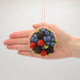 Round hand-felted wool ornament designed as a mixed berry tart, topped with sparkling beaded raspberries, blackberries, and nestled blueberries. Crafted with detailed stitching and vibrant colors, it includes a hanging string and measures approximately 1.25 inches high and 2.75 inches in diameter. A whimsical and delicious-looking tribute to homemade desserts and holiday cheer. Held in a hand for scale.