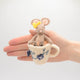 Hand-felted wool ornament of a small gray mouse with a bow, sitting inside a floral teacup with embroidered details. Crafted from merino-blend wool, this whimsical piece evokes tea time and childhood storytelling. Includes a hanging string and measures approximately 4.5 inches tall. A sweet and cozy decoration for tea lovers and mouse enthusiasts alike. Held in a hand for scale. 