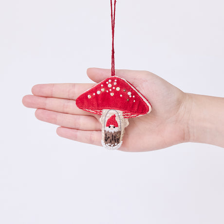 Craftspring’s Mushroom Beaded Ornament held in front of a hand to show its size—handmade from satin and velvet with intricate beaded and embroidered details, including a gnome peeking out of a door on one side of the mushroom, ethically crafted by artisans using sustainable materials.