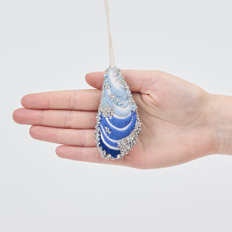 Craftspring’s Mussel Shell Ornament displayed in front of a hand to show its size — made of satin with intricate beaded and embroidered details, ethically crafted by artisans.