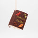 Miniature hand-sewn felt book ornament titled Nature Guide, featuring embroidered and appliquéd illustrations of plants, animals, and woodland scenes. Made from merino-blend wool with whimsical details and a hanging string. Measures approximately 2.5 inches tall by 3 inches wide. A charming tribute to the outdoors and nature lovers.
