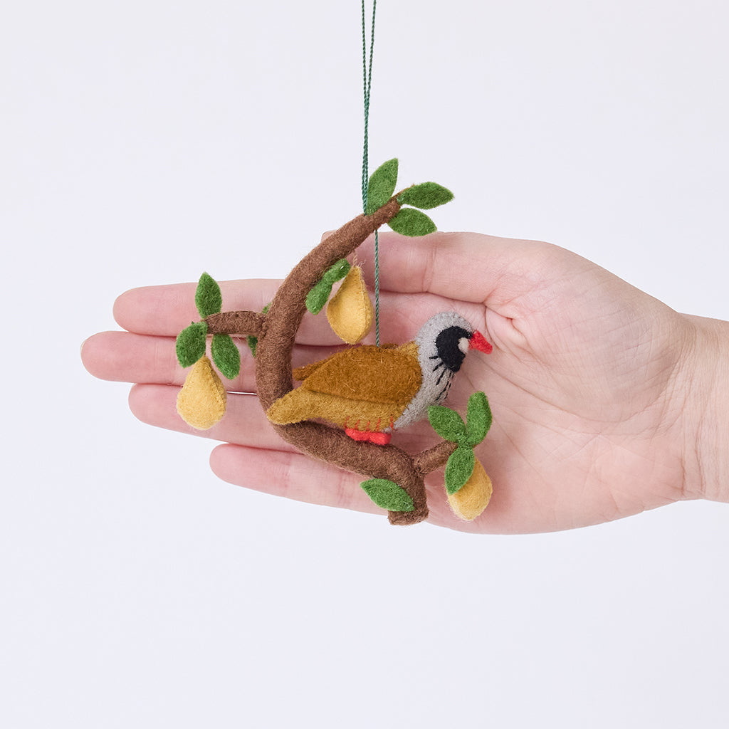Craftspring’s Partridge in a Pear Tree Ornament displayed in front of a hand to show its size — handmade from sustainable wool with embroidered and beaded details of a partridge in a tree with three dangling pears, ethically crafted by artisans.