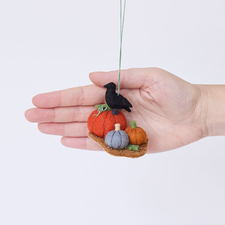 Craftspring’s Pumpkin Patch ornament held in front of a hand to show its size—handmade from sustainable wool with three pumpkins in different colors and a crow perched on top, with beaded and embroidered details, ethically crafted by artisans using sustainable wool.