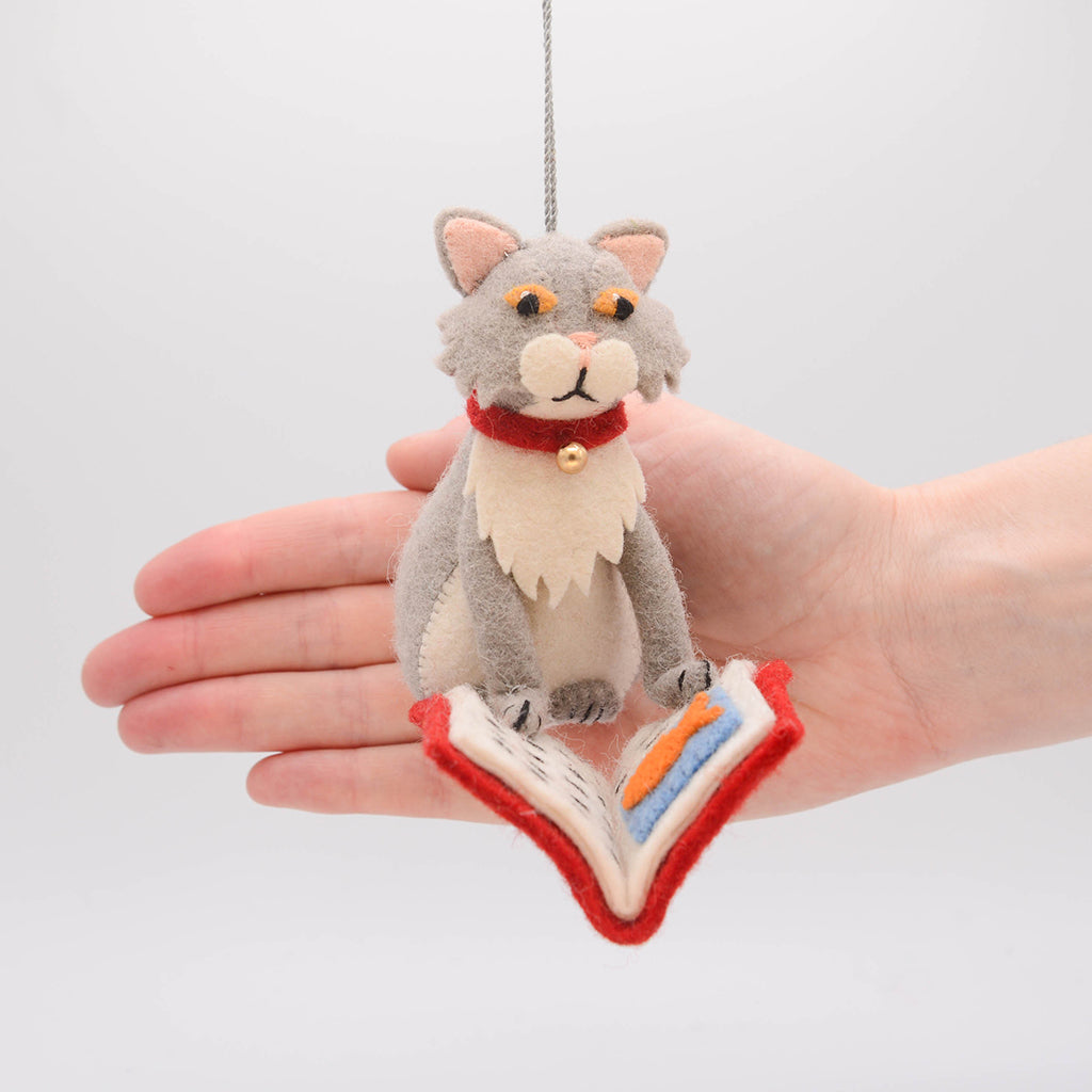 Handmade gray felted cat ornament, depicted mid-chapter in a fishy tale. The playful kitty wears a jingly collar and is fully immersed in its literary adventure. Crafted from natural wool, this whimsical ornament celebrates the love of books and cats, perfect for cat lovers, readers, and bookshop fans. Comes ready to hang with a string, adding a charming touch to any tree or reading nook.