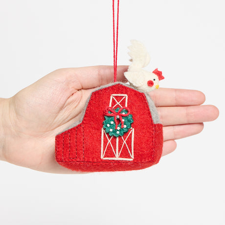 Craftspring’s Red Barn with Rooster Ornament from the Comfort and Joy Ornaments Bundle displayed in front of hand to show its size — felt barn with a rooster on the roof and intricate beaded and embroidered details, a charming heirloom-quality piece for Christmas gifting — ethically crafted by artisans using sustainable wool.