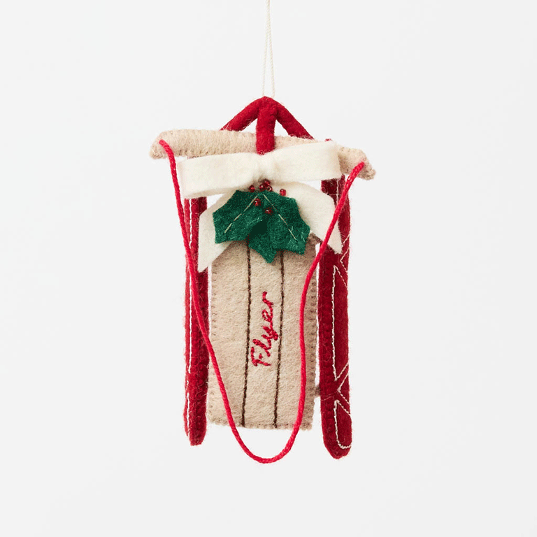 Our Red Flyer Sled Ornament captures the nostalgia of childhood winters and treasured holiday traditions. Inspired by classic wooden sleds, this charming keepsake features bright red runners, a cozy neutral-toned seat embroidered with "Flyer," and a festive holly sprig nestled beneath a soft white bow.