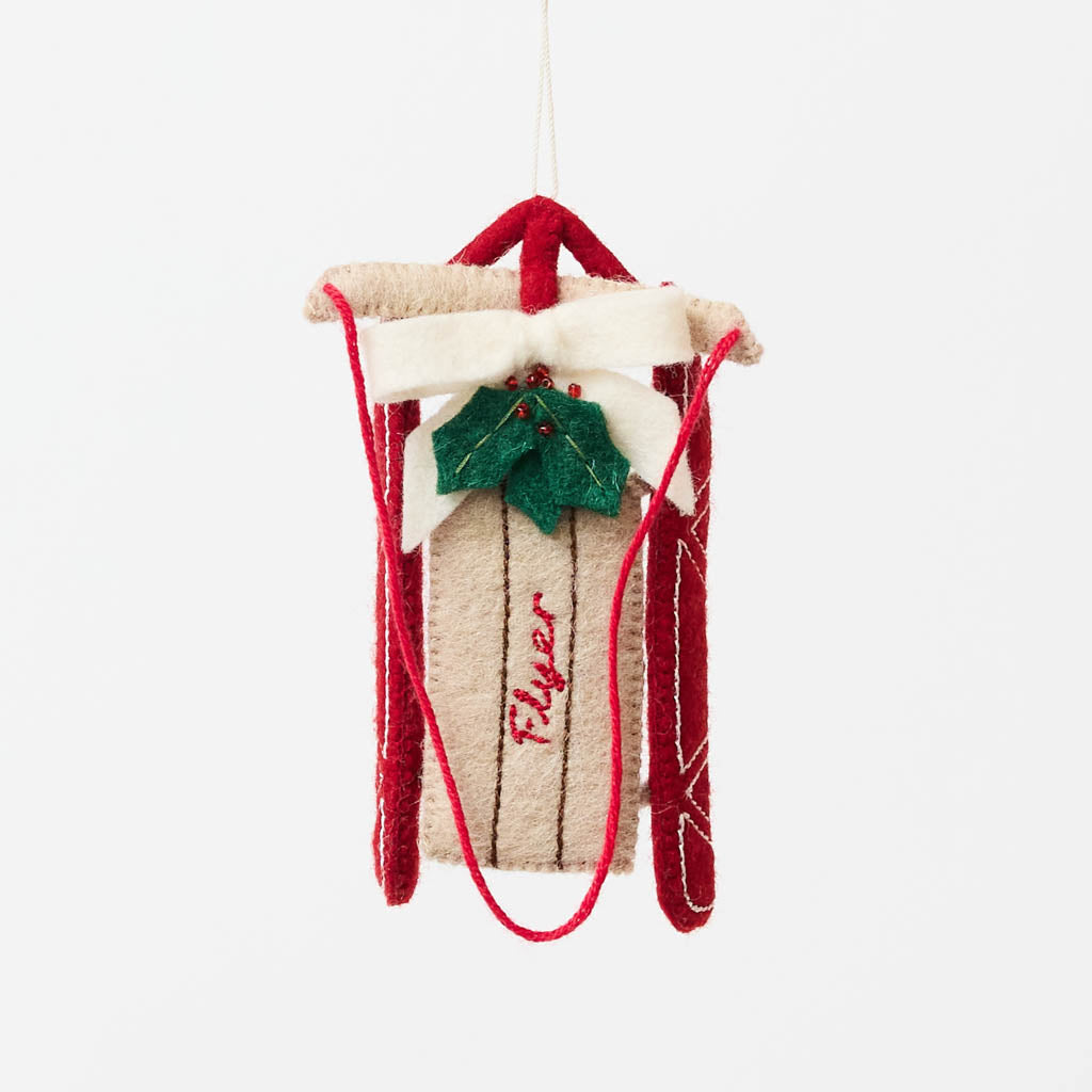 Our Red Flyer Sled Ornament captures the nostalgia of childhood winters and treasured holiday traditions. Inspired by classic wooden sleds, this charming keepsake features bright red runners, a cozy neutral-toned seat embroidered with "Flyer," and a festive holly sprig nestled beneath a soft white bow.