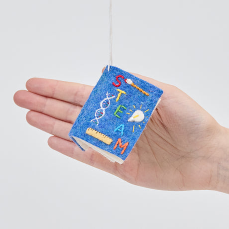 Craftspring’s STEAM Book ornament held in front of a hand to show its size—handmade from sustainable wool with embroidered cover details featuring a DNA strand, paintbrush, light bulb, and ruler, ethically crafted by artisans using sustainable wool.