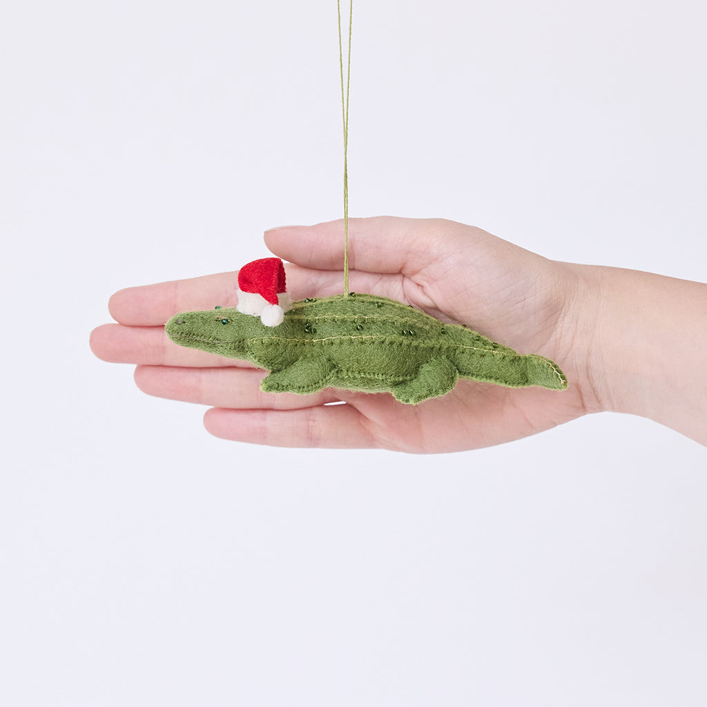 Craftspring’s Santa Alligator Ornament displayed in front of hand to show its size — felt alligator with a beautiful face and embroidered and beaded back ridges, a charming heirloom-quality piece for Christmas gifting — ethically crafted by artisans using sustainable wool.