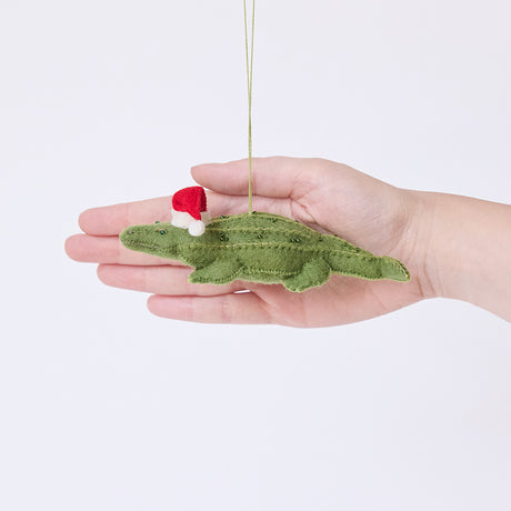 Craftspring’s Santa Alligator Ornament displayed in front of hand to show its size — felt alligator with a beautiful face and embroidered and beaded back ridges, a charming heirloom-quality piece for Christmas gifting — ethically crafted by artisans using sustainable wool.