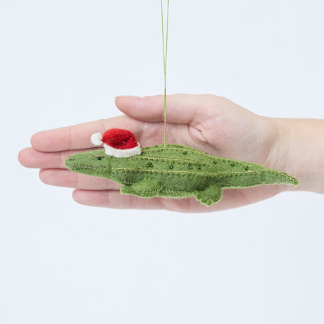 Craftspring’s Santa Alligator Ornament displayed in front of hand to show its size — felt alligator with a beautiful face and embroidered and beaded back ridges, a charming heirloom-quality piece for Christmas gifting — ethically crafted by artisans using sustainable wool.