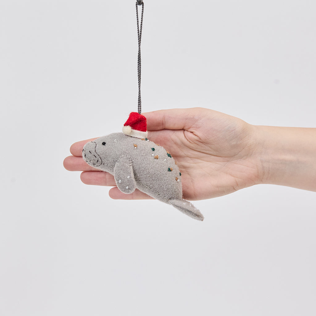 Handmade felt Santa Manatee ornament from Craftspring held in front of a hand to show its size—featuring a red Santa hat and intricate beaded and embroidered details, ethically crafted by artisans using sustainable wool.