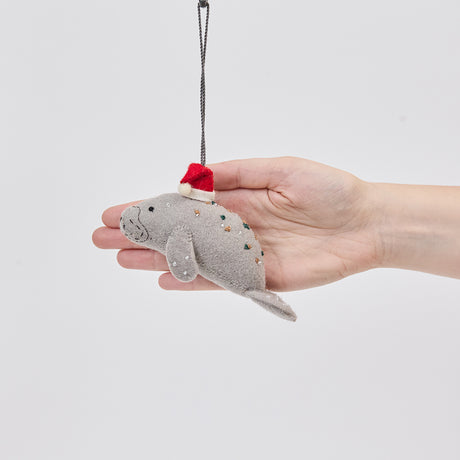 Handmade felt Santa Manatee ornament from Craftspring held in front of a hand to show its size—featuring a red Santa hat and intricate beaded and embroidered details, ethically crafted by artisans using sustainable wool.