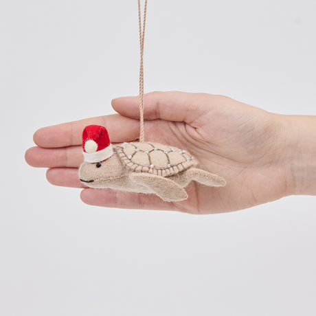 Craftspring’s Santa Neutral Sea Turtle ornament held in front of a hand to show its size—handmade from sustainable wool with intricate beaded and embroidered details and a red Santa hat, ethically crafted by artisans using sustainable wool.