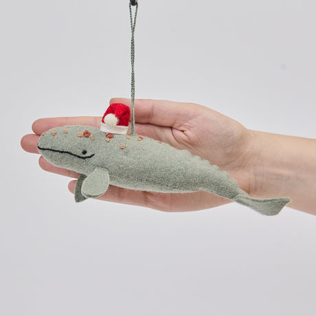 Craftspring’s Santa Spyhop Gray Whale ornament held in front of a hand to show its size—handmade from sustainable wool with intricate embroidered details and a red Santa hat, ethically crafted by artisans using sustainable wool.