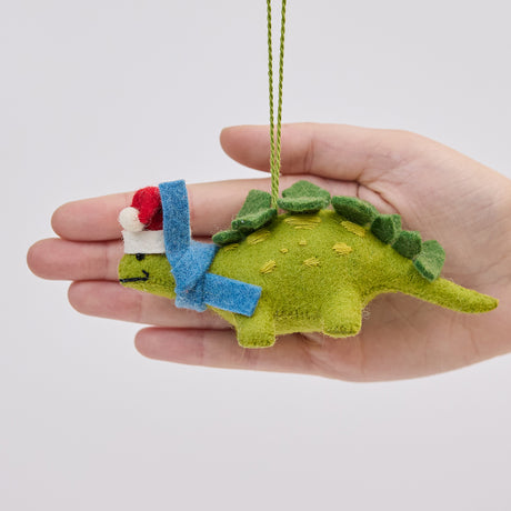 Handmade felt Santa Stegosaurus ornament from Craftspring held in front of a hand to show its size—featuring a green dinosaur with embroidered spots, a red Santa hat, and a blue scarf, ethically crafted by artisans using sustainable wool for whimsical holiday decor.
