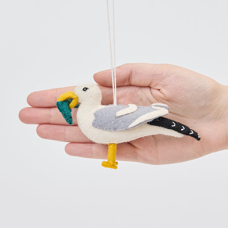 Handmade felt Seagull ornament from Craftspring held in front of a hand to show its size—featuring embroidered details and a fish in its beak, sustainably handmade by artisans using wool.