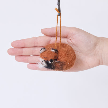 Craftspring’s Sleepy Fox Ornament displayed in front of a hand to show its size — curled up with a cute face, fluffy tail, and whiskers, a sweet heirloom-quality piece perfect for Christmas gifting — ethically crafted by artisans using sustainable wool.