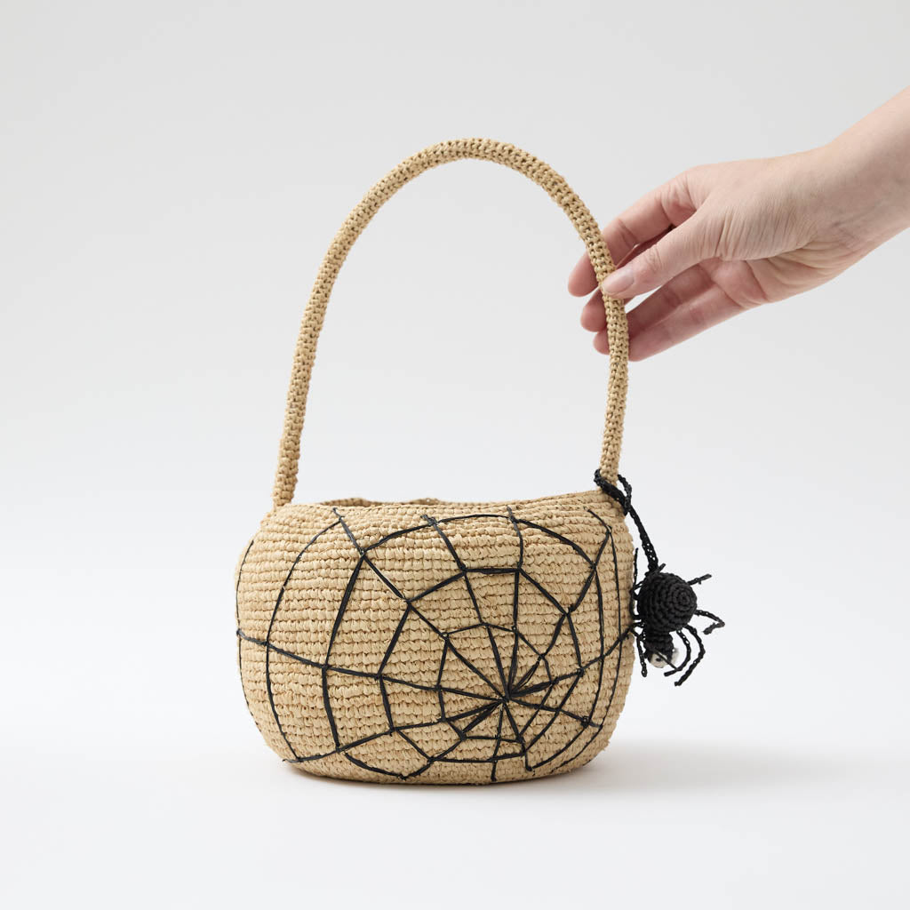 A hand-crocheted raffia Spider Basket featuring a bold black spiderweb and playful spider detail, perfectly sized for trick-or-treating, Halloween treasures, and festive fall displays.