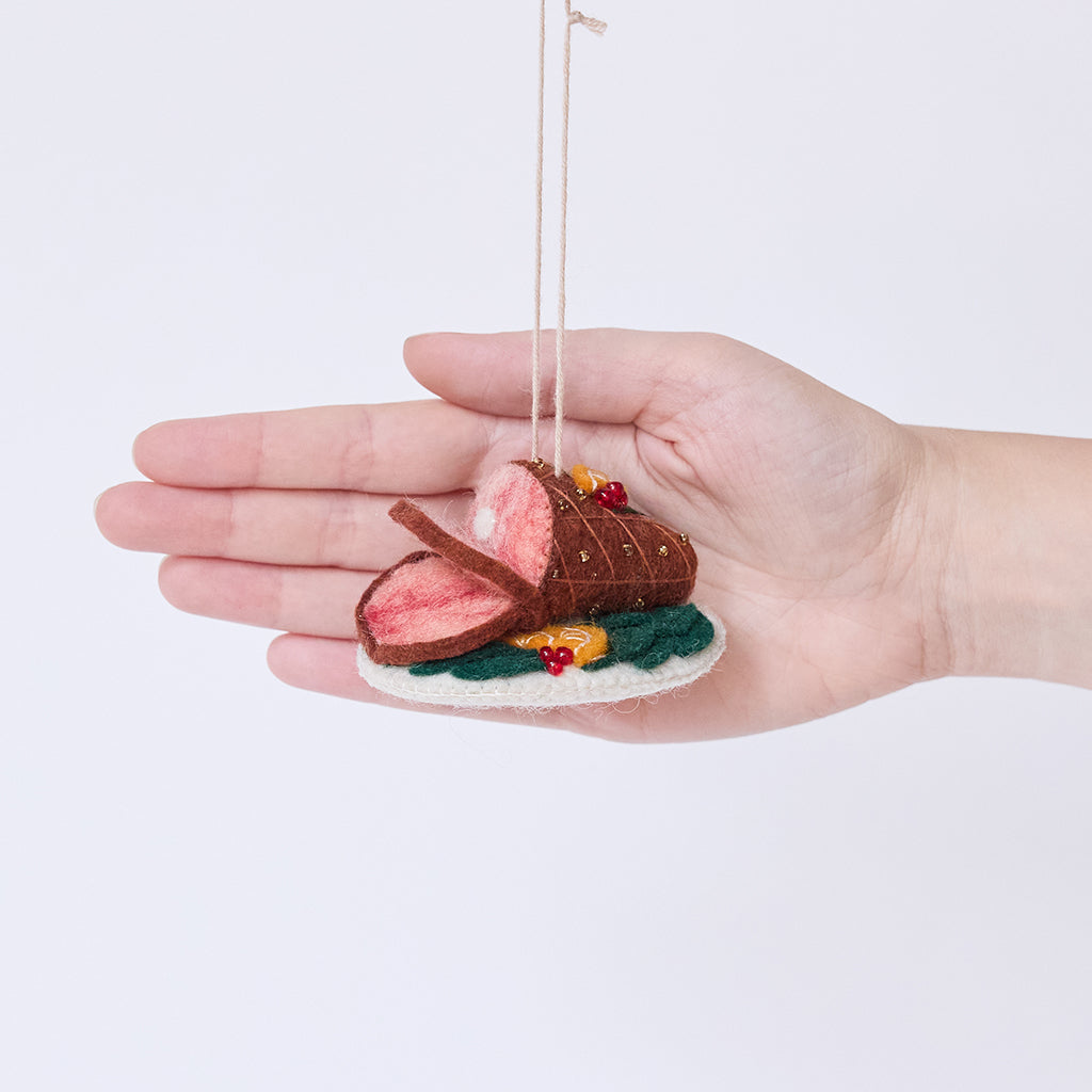 Craftspring’s Spiral Ham Platter ornament held in front of a hand to show its size—handmade from sustainable wool with intricate beaded and embroidered details on a festive ham, ethically crafted by artisans using sustainable wool.