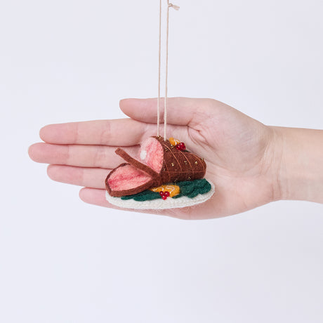 Craftspring’s Spiral Ham Platter ornament held in front of a hand to show its size—handmade from sustainable wool with intricate beaded and embroidered details on a festive ham, ethically crafted by artisans using sustainable wool.