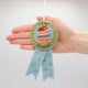 Handmade felt reading badge shaped like a ribbon with shimmering star details, designed to celebrate young readers' achievements. Crafted from merino-blended wool, the badge comes pinned to an illustrated postcard with space for a personal message, and includes an envelope for gifting. Features both a safety pin and a hanging loop. A fun, motivational keepsake for children, perfect for classrooms, libraries, or reading milestones.  With hand for size scale comparison