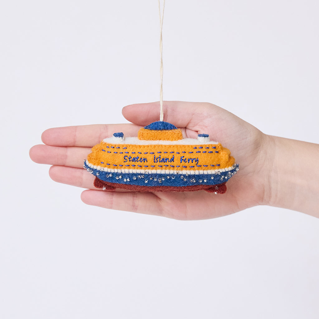 Craftspring’s Staten Island Ferry ornament held in front of a hand to show its size—handmade from sustainable wool with intricate embroidered details and beaded waves, featuring the iconic orange ferry, ethically crafted by artisans using sustainable wool.