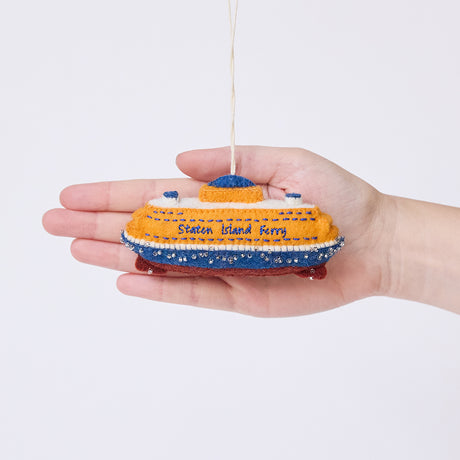 Craftspring’s Staten Island Ferry ornament held in front of a hand to show its size—handmade from sustainable wool with intricate embroidered details and beaded waves, featuring the iconic orange ferry, ethically crafted by artisans using sustainable wool.