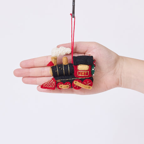 Craftspring’s Steam Engine Train Ornament displayed in front of a hand to show its size — felt black and red train with gold embroidery and beading, fluffy steam, and a wreath on the back, a charming heirloom-quality piece for Christmas gifting — ethically crafted by artisans using sustainable wool.