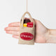 Handmade felt ornament shaped like The Strand Bookstore tote bag, complete with the embroidered red logo and a tiny felt NYC guidebook tucked inside. Crafted from soft tan wool felt, this miniature tote is filled with colorful hand-felted books, paying tribute to the legendary NYC bookstore. A perfect gift for Strand fans, NYC lovers, and book enthusiasts, adding a literary touch to holiday trees or bookshelves.  With a hand of scale comparison.