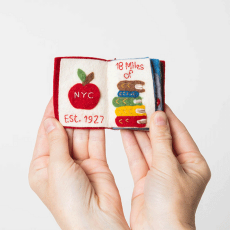Handmade felt Strand Bookstore Book ornament from Craftspring shown flipping through interior pages—featuring intricate beaded and embroidered details inspired by the iconic bookstore, ethically crafted by artisans using sustainable wool.