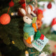 Handmade wool ornament of Peter Rabbit, featuring the beloved children's book character sitting with a carrot in one paw and an embroidered book on his lap. He wears his signature blue jacket, crafted with detailed stitching. Made from natural felted merino-blend wool, the ornament is ready to hang and sized approximately 4.5 inches tall. On a Christmas tree. 