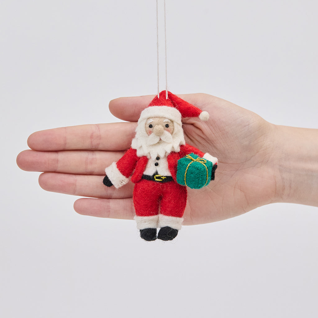 Handmade felt Sweet Little Santa ornament from Craftspring held in front of a hand to show its size—featuring rosy cheeks, a cute round nose, and holding a gift, ethically crafted by artisans using sustainable wool.