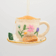Hand-felted ornament of a floral teacup with a matching saucer, featuring embroidered golden rims, a tiny felt tea bag, and two sugar biscuits on the side. Crafted from merino-blend wool with fine needlework, the ornament includes a hanging string and measures approximately 1.5 inches high and 3.25 inches wide. A charming tribute to afternoon tea and cozy traditions. GIF 30 degree view.