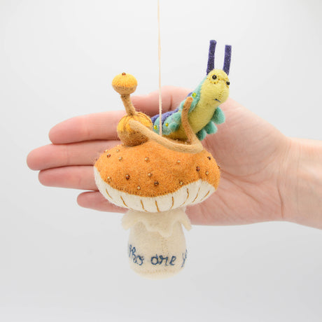 Hand-felted wool ornament of the Caterpillar from Alice in Wonderland, perched on a red-and-white mushroom with a hookah beside him. Features detailed embroidery and the phrase 'Who are you?' stitched at the base. Made from natural merino-blend wool, this whimsical ornament includes a hanging string and measures approximately 5.5 inches tall.
