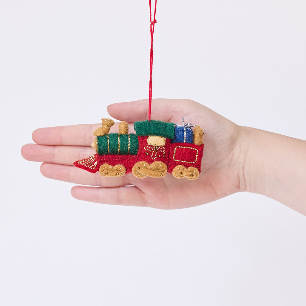 Craftspring’s Toy Train Ornament displayed in front of a hand to show its size — felt red and green train with gold embroidery, carrying presents and a teddy bear, a charming heirloom-quality piece for Christmas gifting — ethically crafted by artisans using sustainable wool.