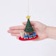 Craftspring’s Train Around the Christmas Tree Ornament displayed in front of a hand to show its size — felt blue toy train wrapping around the base of a decorated Christmas tree with presents, a charming heirloom-quality piece for Christmas gifting — ethically crafted by artisans using sustainable wool.