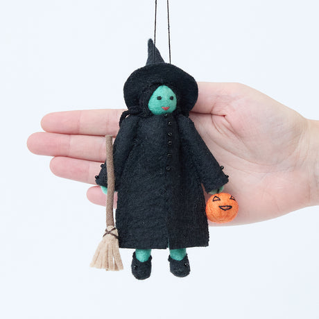 Craftspring’s Wicked Witch Trick-or-Treater Ornament displayed in front of a hand to show its size—handmade felt witch with green skin, hat, broom, and pumpkin basket, heirloom-quality Halloween decor, perfect for holiday gifting, ethically crafted by artisans using sustainable wool.