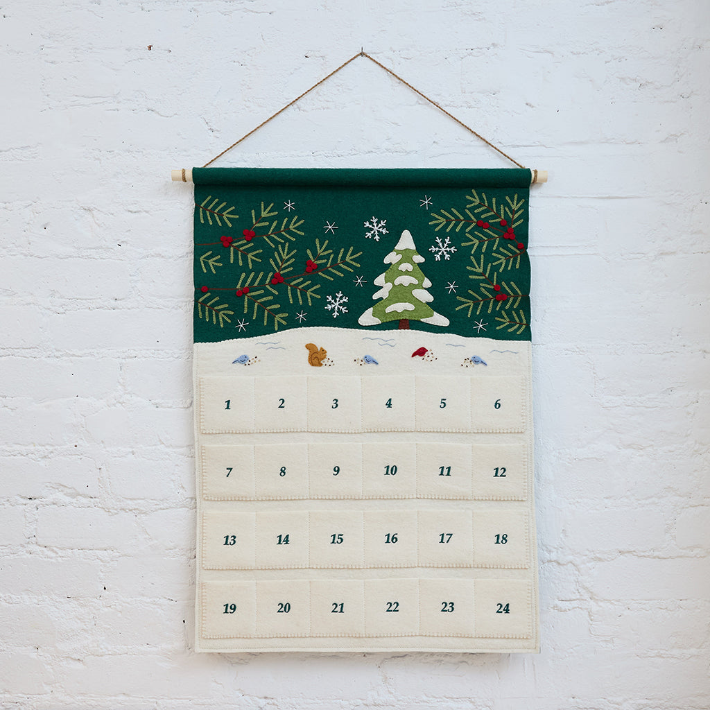 Craftspring’s Woodland Advent Calendar — handmade felt calendar with appliqué, embroidered, and beaded details of a snowy woodland scene featuring a snowy tree, birds, a squirrel, evergreen branches with red berries, and snowflakes — heirloom-quality Christmas decor, ethically crafted by artisans using sustainable wool.