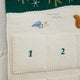 Craftspring’s Woodland Advent Calendar — handmade felt calendar with appliqué, embroidered, and beaded details of a snowy woodland scene featuring a snowy tree, birds, a squirrel, evergreen branches with red berries, and snowflakes — heirloom-quality Christmas decor, ethically crafted by artisans using sustainable wool.