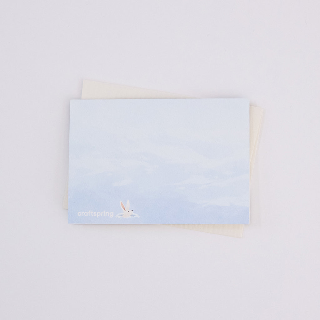 Back of Craftspring’s Woodland Burrows Greeting Card, showing an illustration of a bunny peeking its head out of a hole in the snow, ethically printed in the USA on natural textured paper, perfect for sharing holiday cheer and gifting.