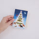 Craftspring’s Woodland Tree Greeting Card held in hand to show its size—featuring woodland animals around a glowing winter Christmas tree, ethically printed in the USA on natural textured paper, perfect for sharing holiday cheer and gifting.