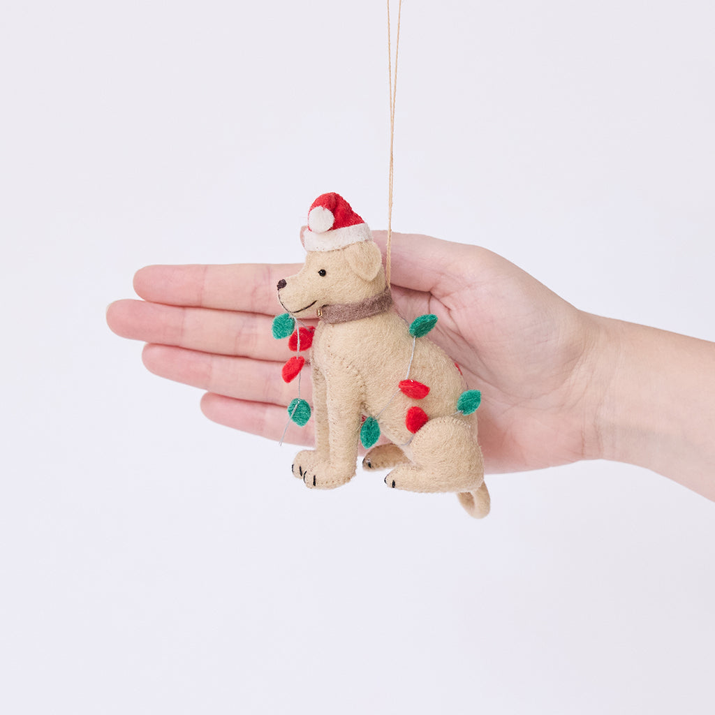 Craftspring’s Yellow Lab in Christmas Lights Ornament held in hand to show its size — felt dog wrapped in Christmas lights and wearing a Santa hat, a charming heirloom-quality piece for Christmas gifting — ethically crafted by artisans using sustainable wool.