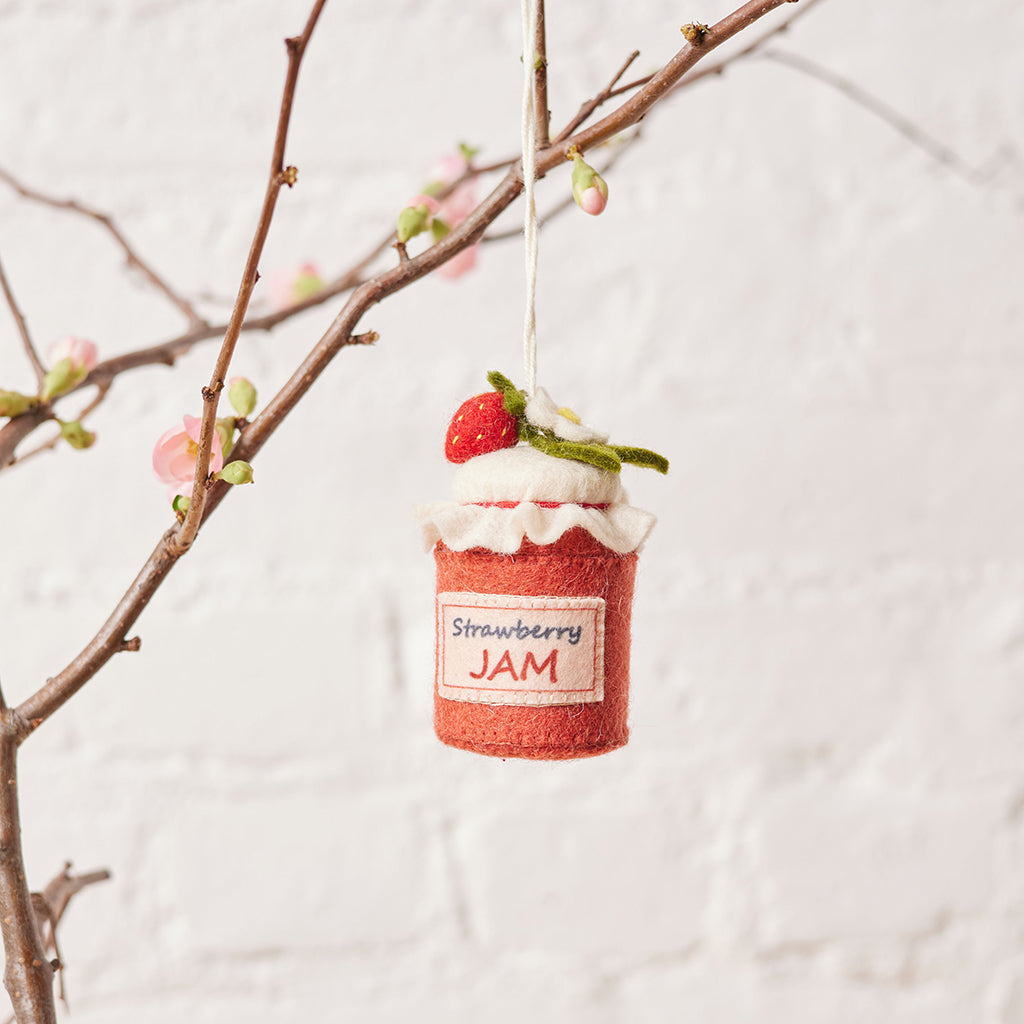 Hand-felted wool ornament of a strawberry jam jar, topped with a white gingham-style lid and a blooming felt strawberry. Features a stitched label and vibrant red detailing, evoking homemade summer preserves. Measures 3.25 inches tall and includes a hanging string — a charming gift for gardeners, bakers, or lovers of nostalgic, handmade summer treasures.