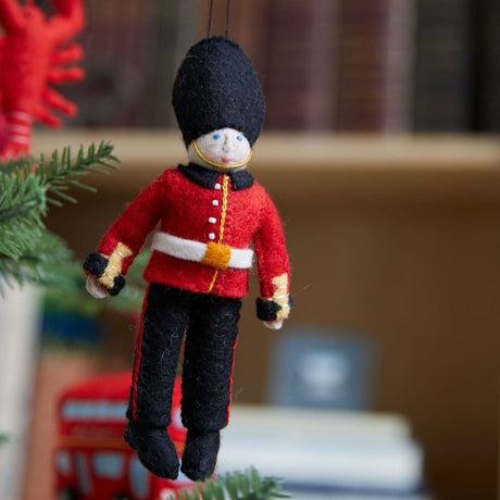 Queen’s Guard felt ornament wearing red coat and black bearskin hat, inspired by Buckingham Palace guards. Handmade wool Christmas ornament celebrating British tradition. Fair trade and ready to hang.
