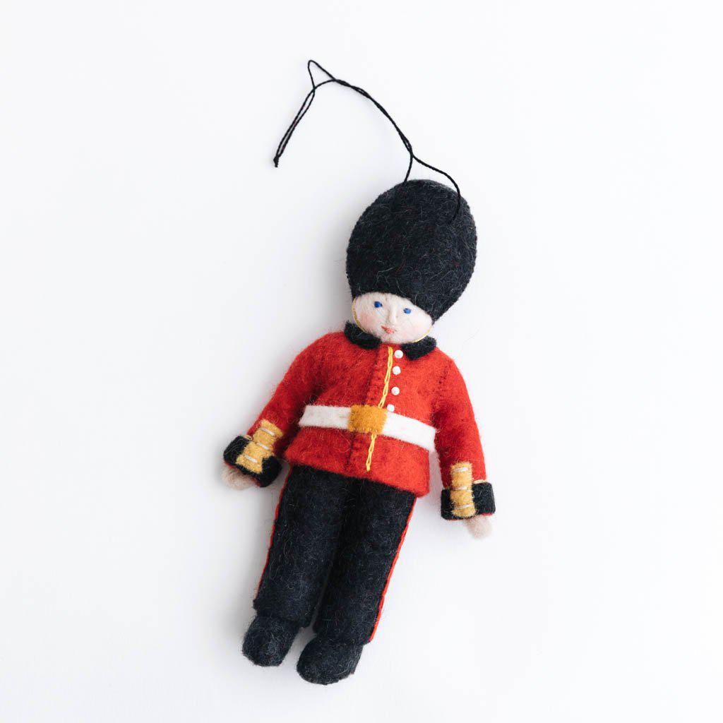 Queen’s Guard felt ornament wearing red coat and black bearskin hat, inspired by Buckingham Palace guards. Handmade wool Christmas ornament celebrating British tradition. Fair trade and ready to hang.